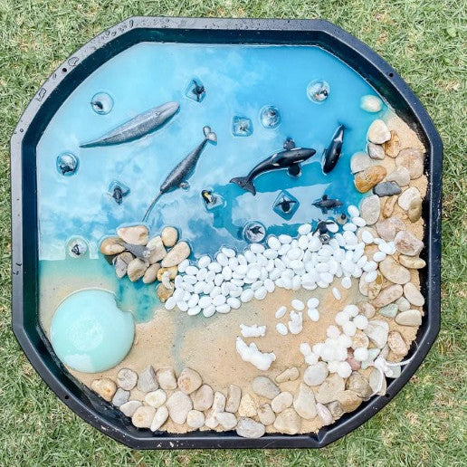 Decorative garden pond with fish figures, pebbles, and a blue water surface on a black tray on grass.
