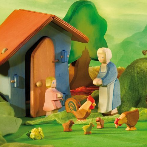 A colorful wooden toy farm scene with a small blue house, orange roof, chickens, a rooster, a woman, a child, and a cow on a green painted backdrop.